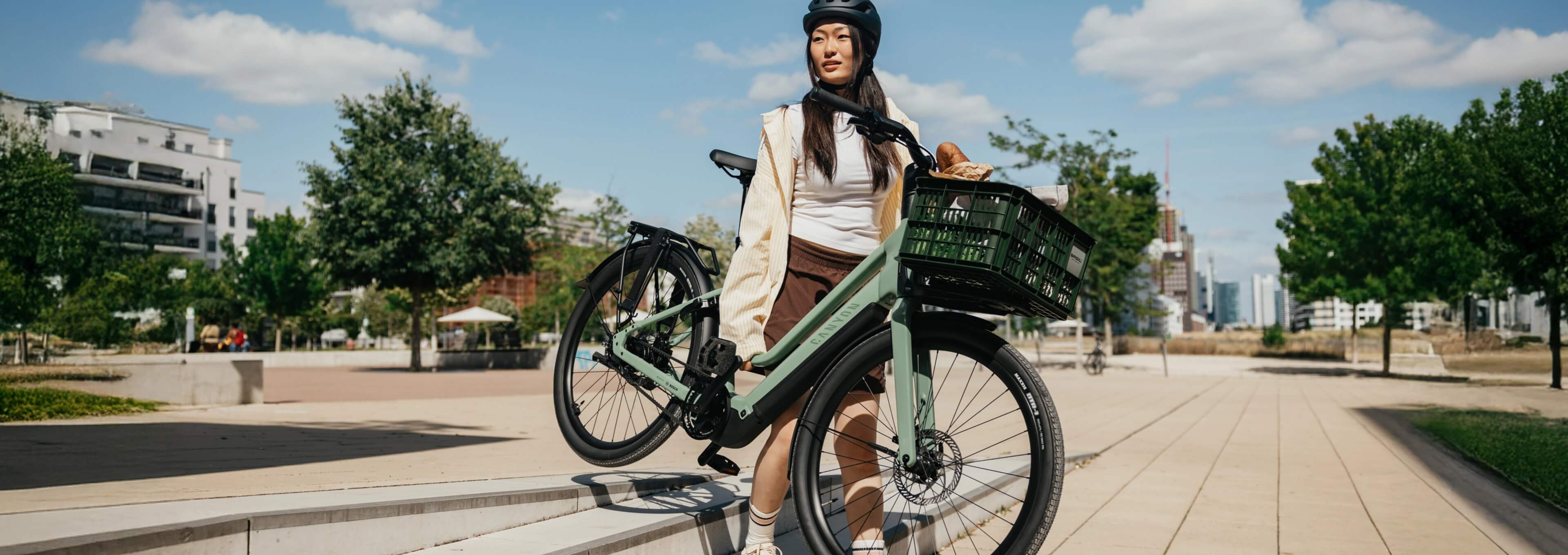 Person mit Helm trägt ein grünes E-Bike mit Frontkorb eine Treppe in einem städtischen Park hinauf.