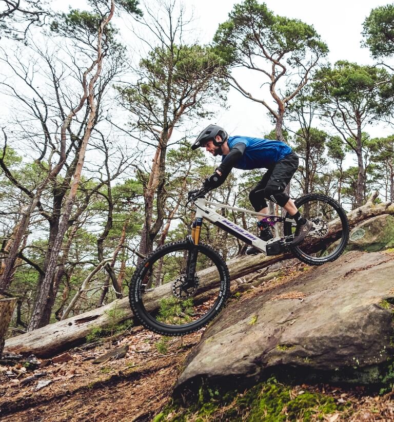 Mountainbiker mit blauer Jacke fährt einen steilen, felsigen Trail im Wald hinab.