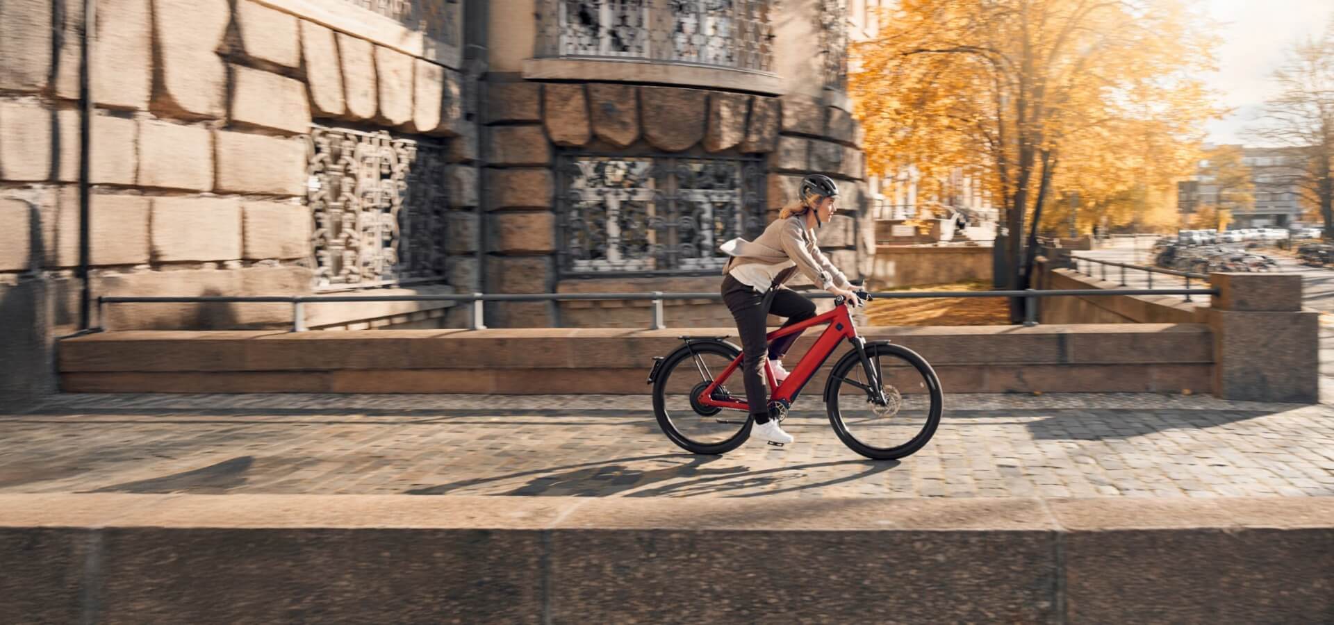 Person fährt auf einem roten Fahrrad eine Stadtstraße entlang, neben einem alten Steingebäude und herbstlichen Bäumen.