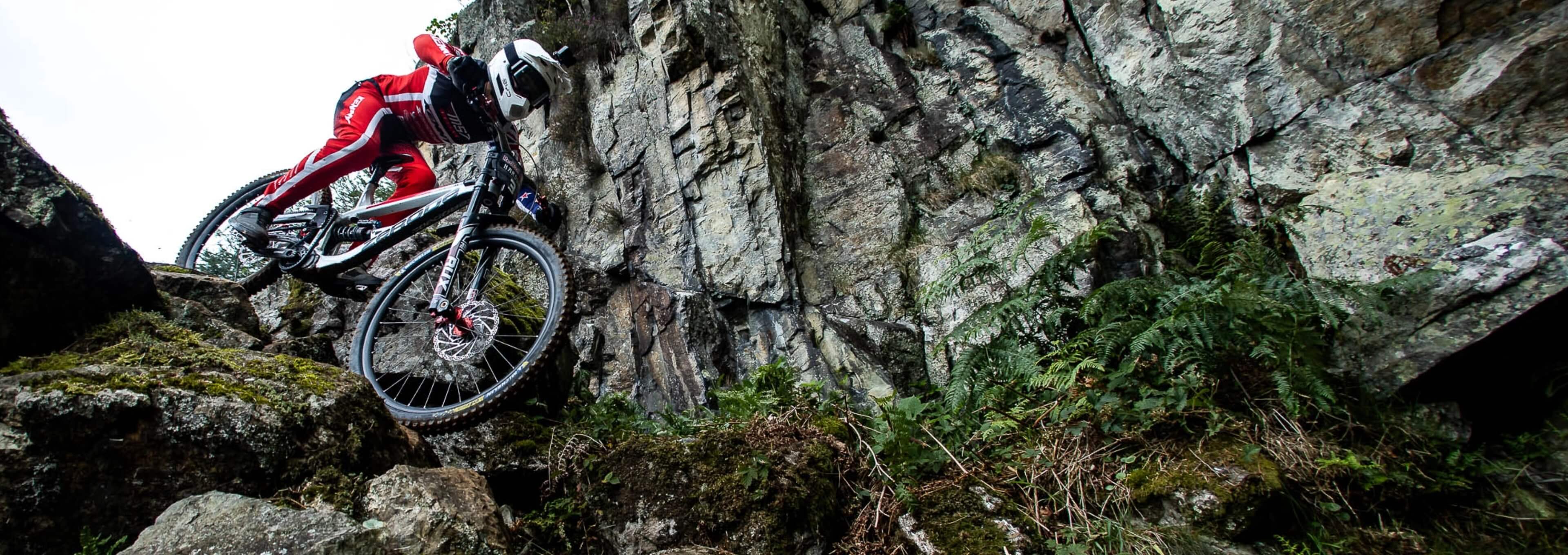 Mountainbiker in roter Kleidung fährt einen steilen, felsigen Trail neben einer Felswand hinab.