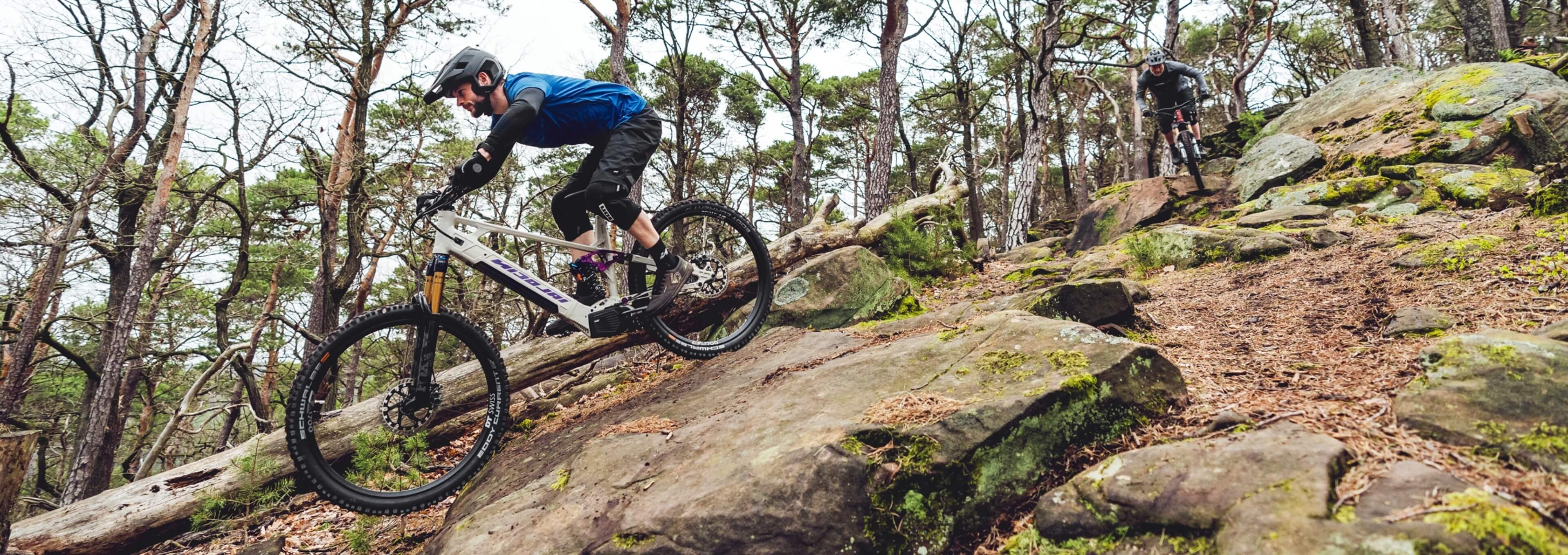 Zwei Mountainbiker fahren einen steilen, felsigen Trail durch den Wald hinab.
