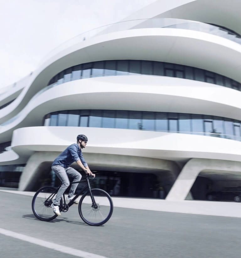 Person fährt mit einem modernen Citybike an einem großen, geschwungenen, futuristischen Gebäude an einem hellen Tag vorbei.