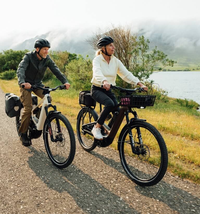 Zwei Personen mit Helmen fahren auf E-Bikes eine Landstraße am See entlang, im Hintergrund liegen nebelverhangene Berge.