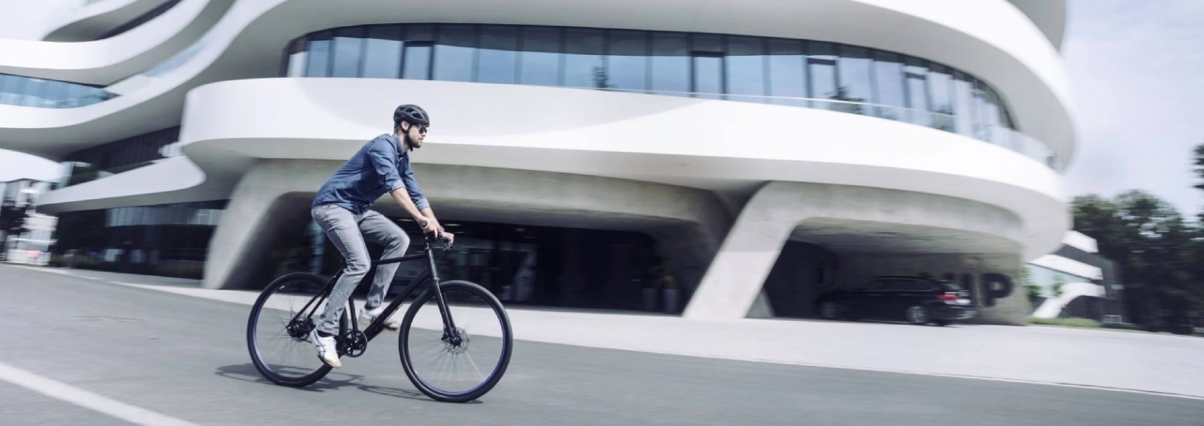 Person fährt mit einem modernen Citybike an einem großen, geschwungenen, futuristischen Gebäude an einem hellen Tag vorbei.