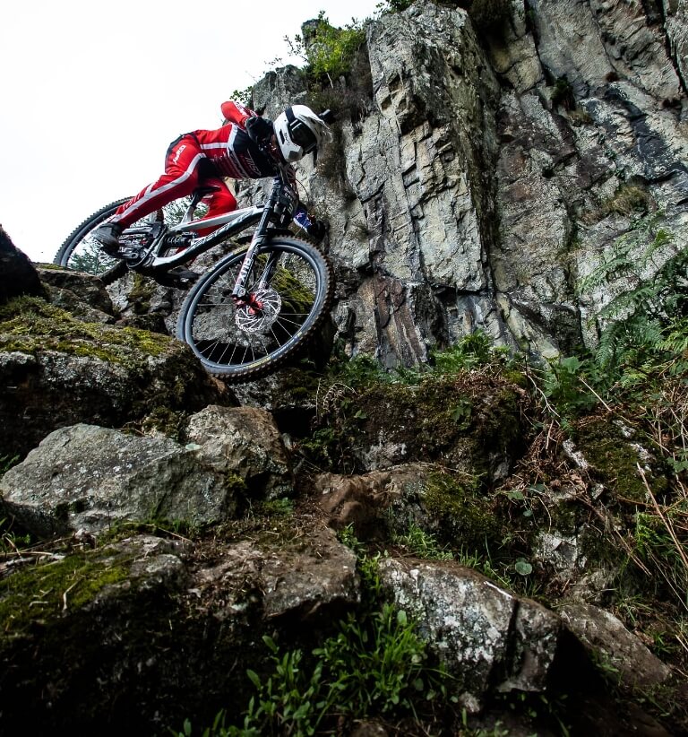 Mountainbiker in roter Kleidung fährt steil einen felsigen Trail an einer Felswand hinab.