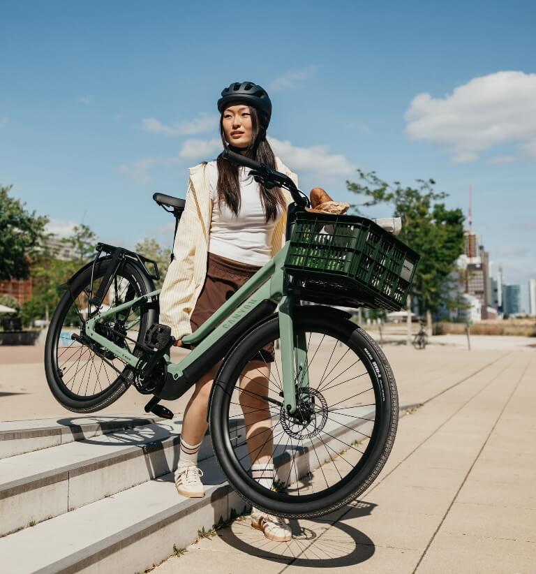 Person mit Helm trägt ein grünes E-Bike mit Frontkorb eine Treppe in einem städtischen Park hinauf.
