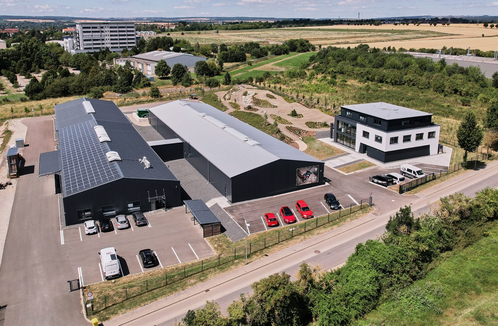 Luftaufnahme eines modernen Industriegeländes mit zwei langgezogenen Lagerhallen, einem Bürogebäude, Parkplätzen mit Autos und umliegenden Grünflächen.