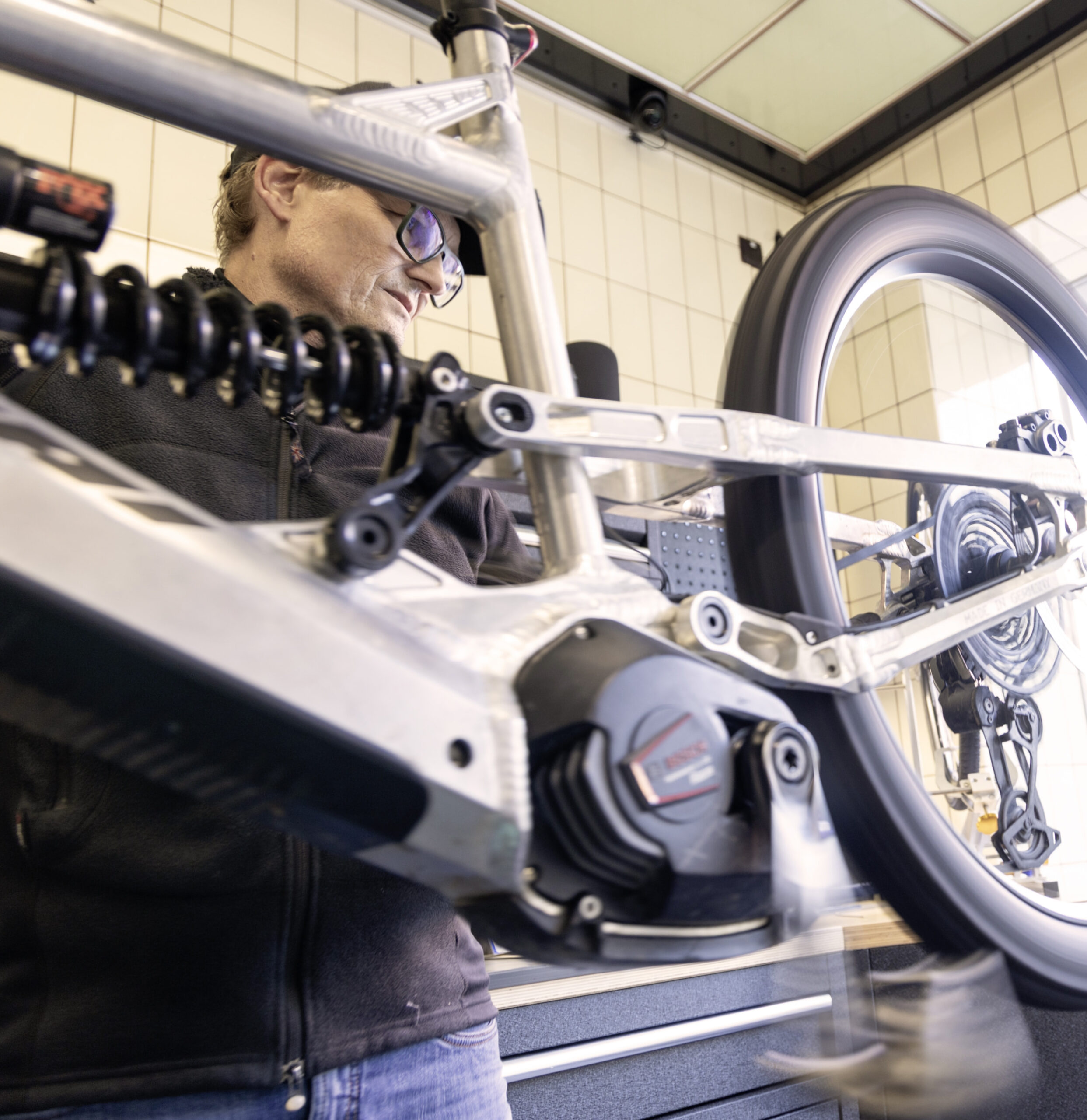 Nahaufnahme eines E-Mountainbikes in der Werkstatt mit Fokus auf Motor und Hinterradfederung, Mechaniker im Hintergrund.